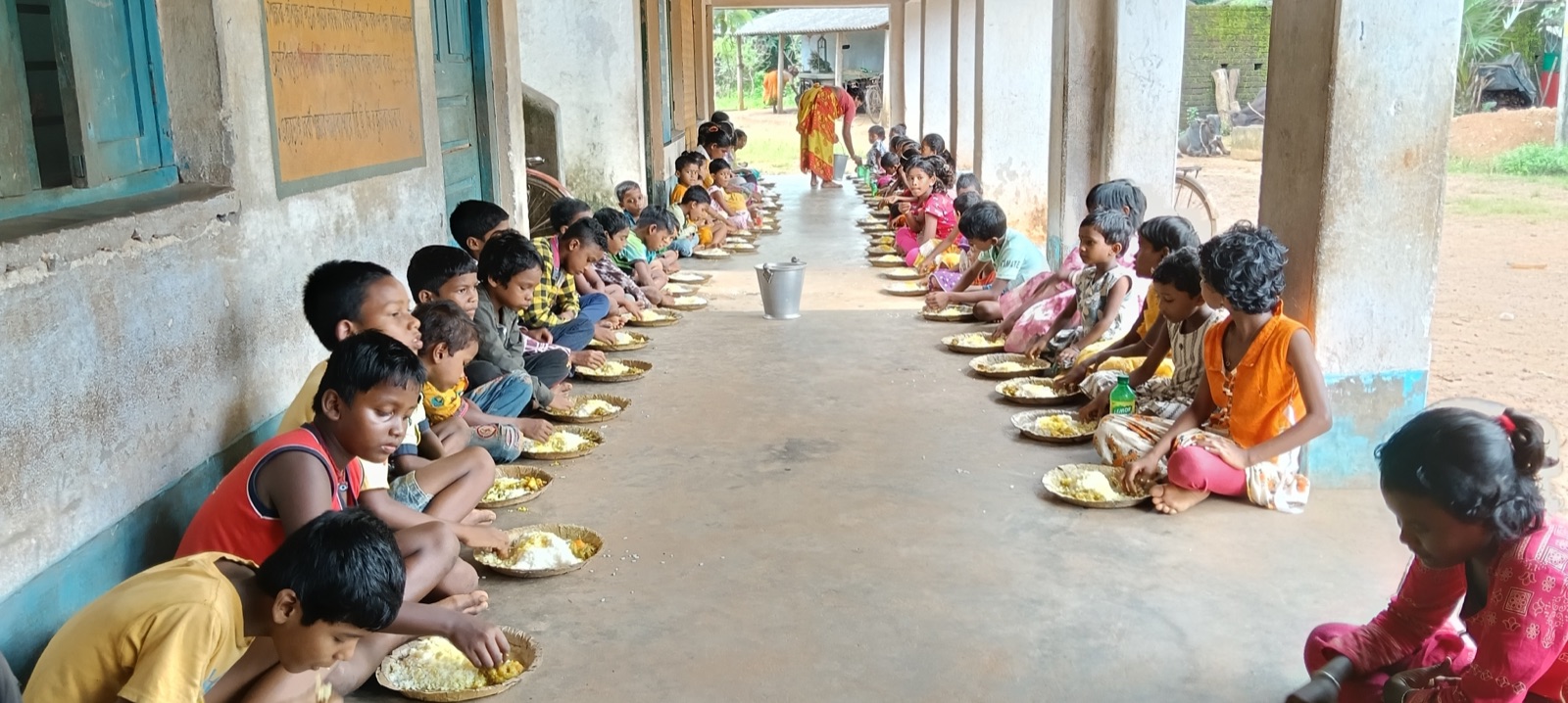 Two long rows of tribal schoolchildren eating a meal along a school veranda in West Bengal, India