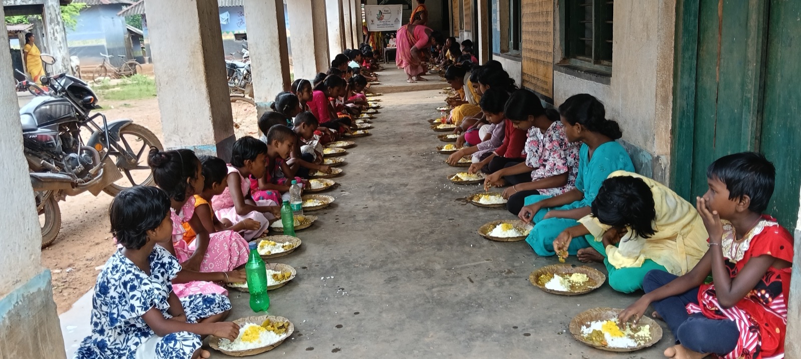 Girls seated along the veranda eating rice, dal, and vegetables from leaf plates